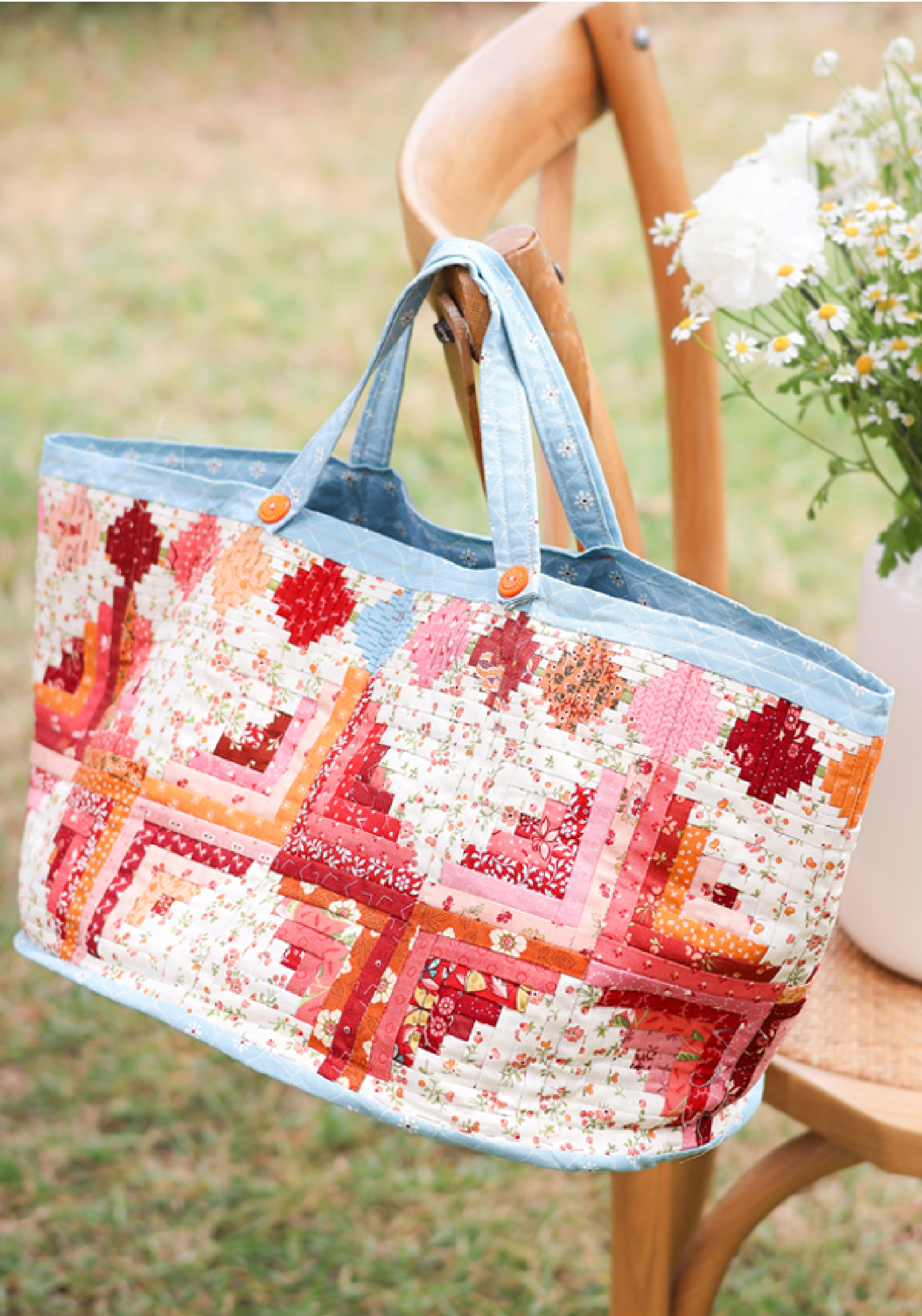 Sac cabas patchwork coloré avec motifs quilt floraux et anses en tissu bleu, suspendu à une chaise en bois près d’un bouquet de fleurs, idéal pour couture DIY, patchwork, quilting et accessoires mode faits main