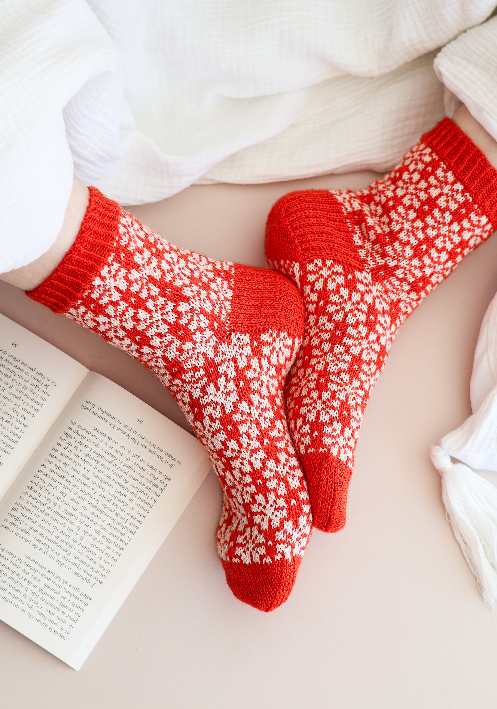 Chaussettes tricotées rouges et blanches à motifs jacquard portées dans une ambiance cocooning avec livre ouvert, idéales pour tricot DIY, accessoires hiver, mode handmade et moments cosy à la maison