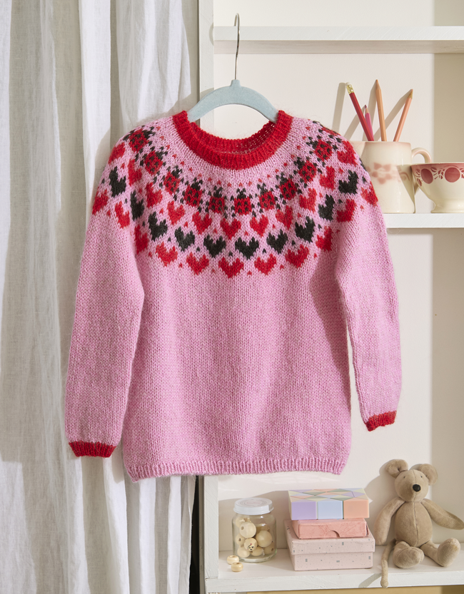 Pull en tricot jacquard rose à motifs rouges, noirs et blancs, présenté sur un cintre.