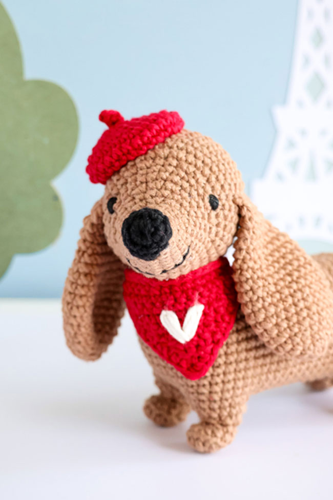 Chien amigurumi crocheté à la main avec béret rouge et foulard assorti en forme de cœur, création artisanale pleine de charme idéale pour crochet DIY, peluche handmade, univers kawaii et loisirs créatifs.