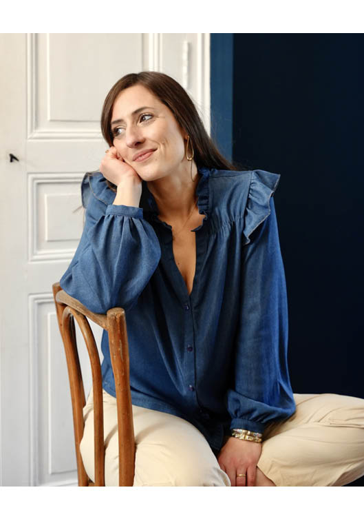 Portrait lifestyle d’une femme brune assise sur une chaise en bois, portant une blouse bleue élégante à volants, ambiance chic et naturelle, idéal pour mode féminine, portrait lifestyle et inspiration style casual élégant