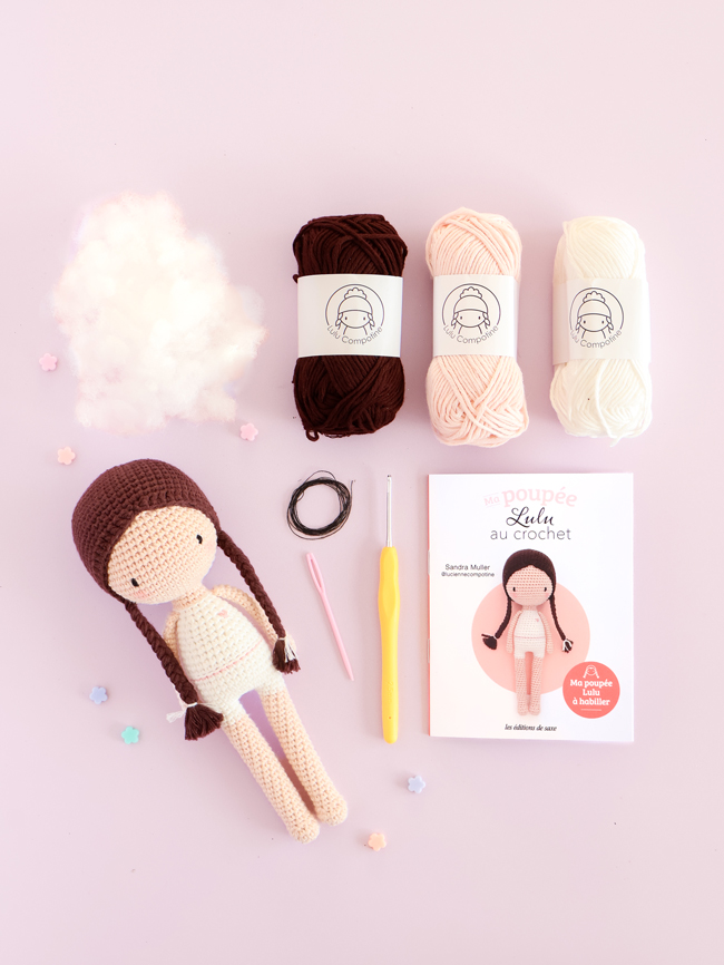 Kit de crochet Ma poupée Lulu avec la poupée à crocheter, trois pelotes de laine, un crochet, une aiguille, du rembourrage, un élastique et le livret d'instructions de Sandra Muller (Lulu Compotine), présenté sur un fond rose pastel.