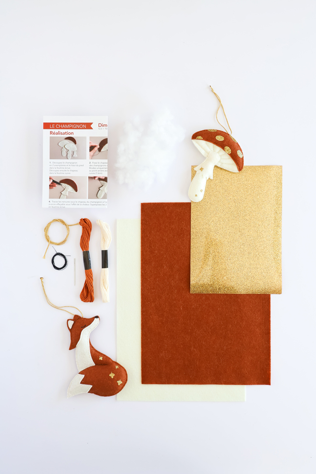 Contenu du kit créatif Champignon : feutrine, tissu pailleté doré, fils à broder, rembourrage, cordon, accessoires de montage, tutoriel pas à pas et suspensions champignon et oie, présenté sur fond blanc.