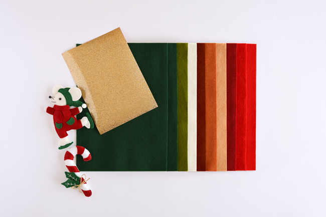 Contenu du kit créatif de Noël : assortiment de feuilles de feutrine aux couleurs de Noël, coupon de tissu pailleté doré et petit lutin décoratif, présenté sur fond blanc.