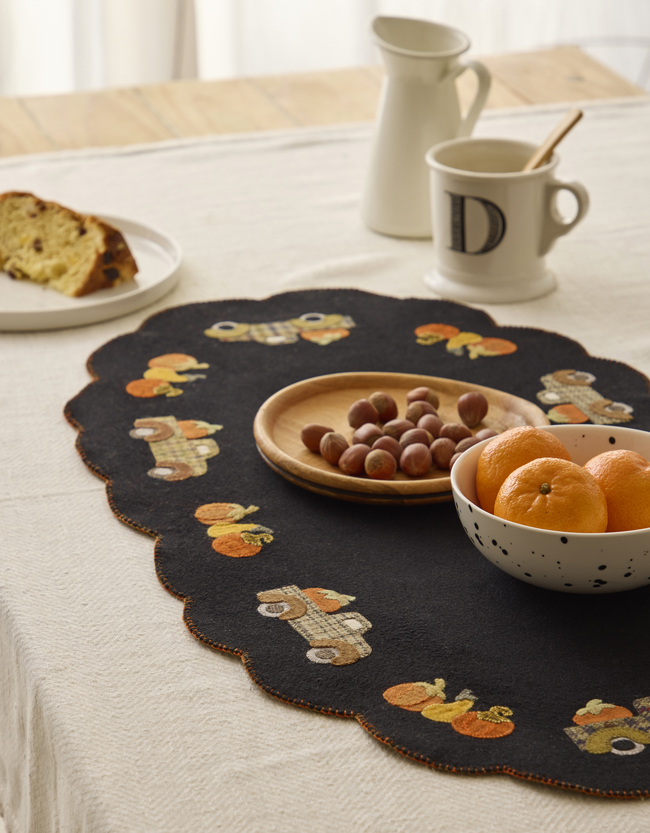 Centre de table en feutrine noire brodé de motifs d’automne avec citrouilles et petits personnages, décoré de fruits de saison.