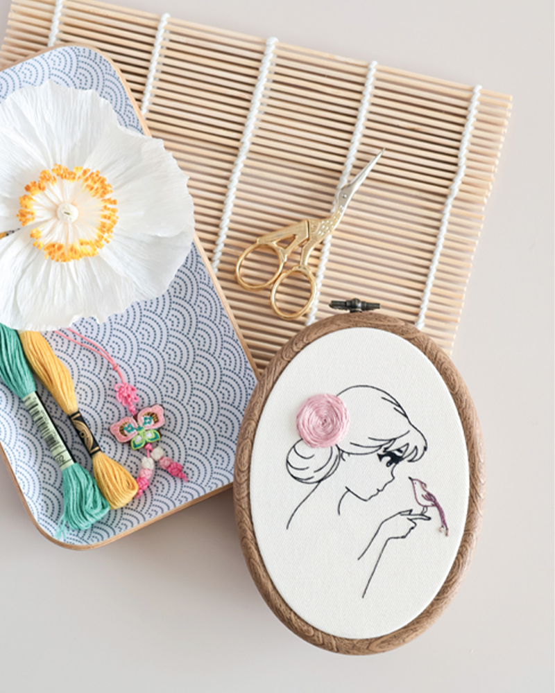 Tambour à broder avec illustration minimaliste d’une femme et détails brodés, entouré de fils à broder, ciseaux et accessoires créatifs, idéal pour broderie DIY, loisirs créatifs, couture décorative et créations textiles faites main.