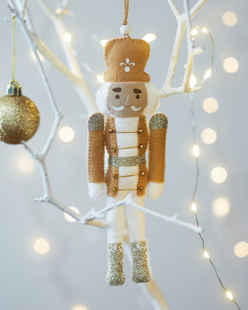 Ornement de Noël casse-noisette en tissu et feutrine aux détails dorés, suspendu à une branche décorée de guirlandes lumineuses, idéal pour décoration de Noël DIY, loisirs créatifs, ambiance festive et créations artisanales faites main.