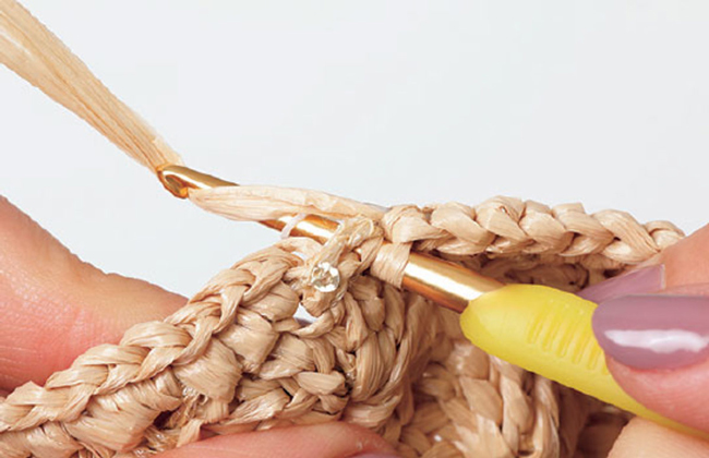 Gros plan d’une étape de crochet en raphia montrant le passage du crochet dans les mailles, idéal pour tutoriel crochet DIY, techniques de crochet, apprentissage des points et réalisation d’accessoires en fibres naturelles.